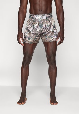 Erwachsener Mann trägt glänzende, camouflagegemusterte Venum-Boxershorts, steht barfuß mit leicht gespreizten Beinen vor einfachem Hintergrund.