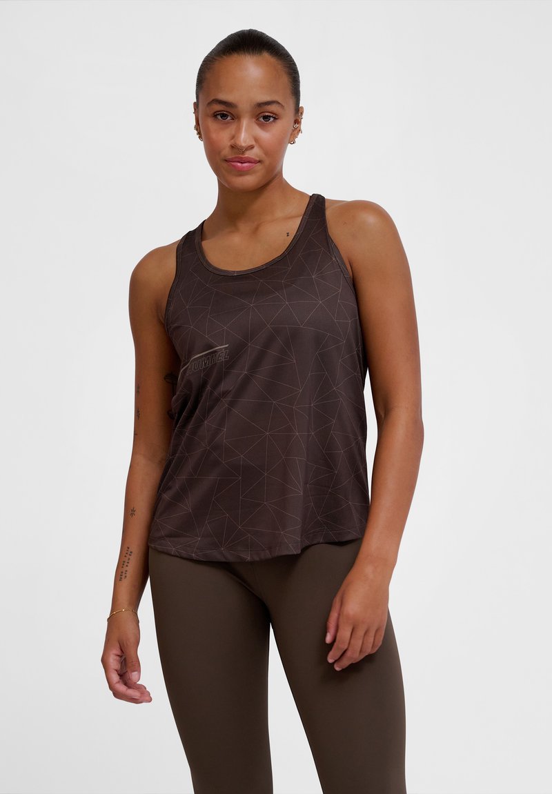 Femme portant un haut de sport sans manches marron foncé à motif géométrique et des leggings assortis, debout devant un fond clair uni.