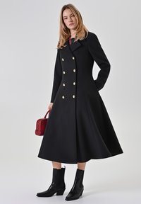 Schwarzer doppelt geknöpfter Mantel mit goldenen Knöpfen, ausgestelltem Rockteil und langen Ärmeln. Kombiniert mit schwarzen Stiefeletten und einer kleinen roten Handtasche.
