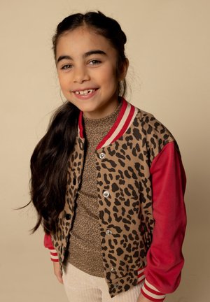 Leopard-print bomberjack met rode mouwen en gestreepte kraag; gedragen boven een beige, textuur top; lang, donker haar met een vlecht.