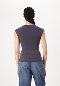 Personne avec des cheveux tressés portant un haut sans manches gris foncé et un jean bleu, se tenant de dos devant un fond blanc uni.