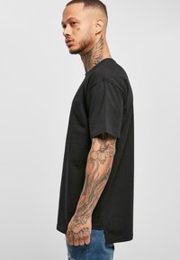 Svart bomulls t-skjorte med korte ermer, rund hals, og sidesplitter, vist på en person med tatoveringer og denimjeans.