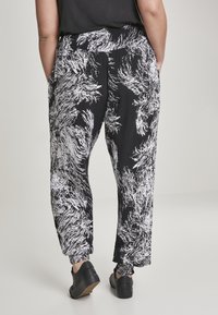 Personne portant un pantalon large noir et blanc à motif avec des poignets élastiques et des chaussures décontractées noires, se tenant avec les mains dans les poches.