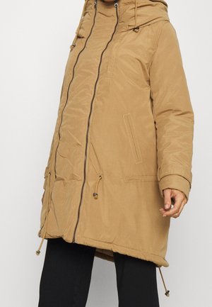 Beżowa długa parka z kapturem, z zamkiem z przodu, bocznymi kieszeniami i regulowanymi troczkami, noszona na czarnych spodniach.