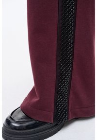 Bordowe spodnie dresowe z czarnym teksturowanym paskiem w romby wzdłuż boku, zestawione z błyszczącym czarnym butem.