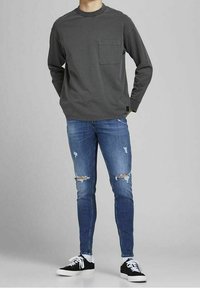 Maglietta grigia a maniche lunghe con taschino sul petto, abbinata a jeans blu strappati con buchi e sneakers nere con lacci bianchi.