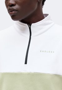 Gros plan d'une personne portant un pull à quart de zip blanc et vert clair avec le texte "ENDLESS" sur la poitrine.