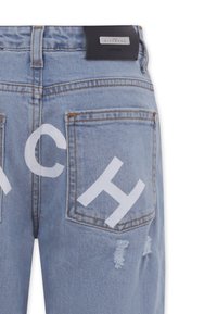 Jeans in denim azzurro chiaro con dettagli strappati, grande branding bianco sulle tasche posteriori e un'etichetta nera con testo in vita.