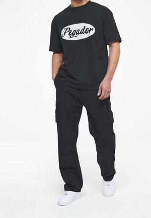 Czarna koszulka z białym logo "Pegador", zestawiona z czarnymi spodniami cargo z kieszeniami oraz białymi trampkami. Prosty, luźny krój.