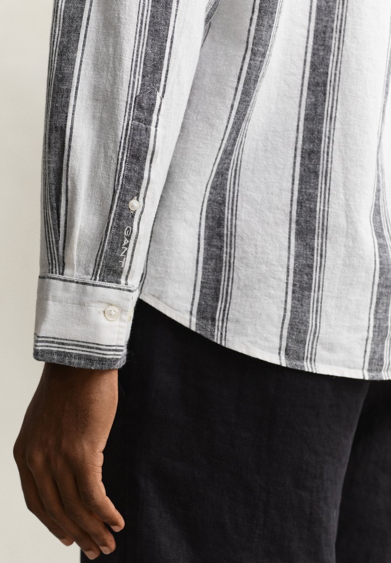 Donkere hand langs het lichaam rustend, met een wit hemd met lange mouwen met zwarte verticale strepen en zwarte broek.