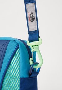 Mochila azul e verde-água com uma superfície texturizada, apresentando um mosquetão verde e um rótulo cinza. Detalhe do fecho éclair e bolso lateral em malha visível.