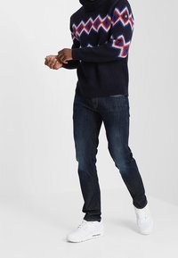 Marinblå högre hals tröja med röd och vit vågmönster, parat med mörkblå jeans och vita sneakers, stående mot en ljus bakgrund.