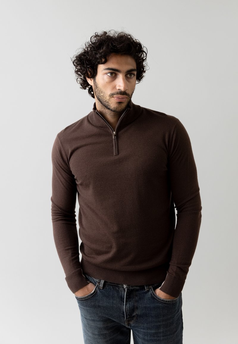 Brauner Zip-Kragen-Pullover mit langen Ärmeln, aus weichem Material. Getragen mit dunkelblauen Jeans. Einfaches, figurbetontes Design. Keine sichtbaren Muster oder Akzente.