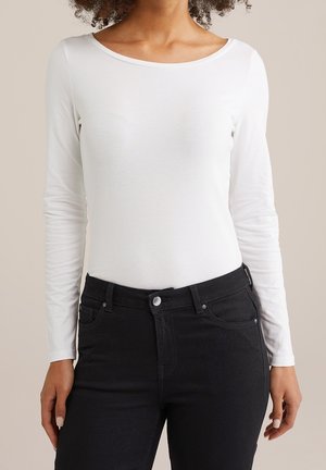 Mujer con una camisa blanca de manga larga lisa metida en unos vaqueros negros de cintura alta, de pie frente a un fondo neutro.