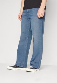 En man i lösa blå jeans och svarta låga sneakers med vita sulor, som står mot en enfärgad ljus bakgrund.