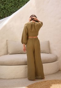 Femme portant un haut à manches longues vert olive et un pantalon large, debout de dos à la caméra, main touchant les cheveux, près d’un banc beige rembourré.