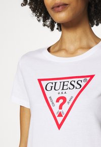 Hvid bomulds t-shirt med et stort rødt trekantet grafisk motiv med sort tekst og et spørgsmålstegn, mærket "GUESS U.S.A. WASHED JEANS."