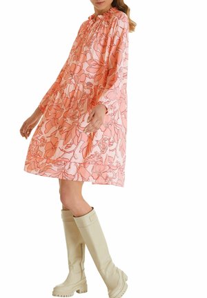 Nainen, joka wear loose pinkki kukkamekko pitkillä hihoilla, yhdistettynä polven ylle ulottuviin valkoisiin paksupohjaisiin saappaisiin, seisoo sivuttain.