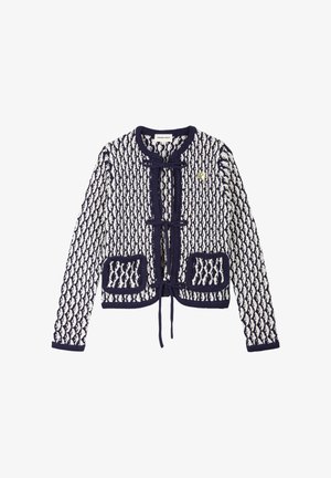 Marineblaue und weiße Strickjacke mit Diamantmuster, vorderen Binderverschlüssen, zwei Vordertaschen und einer kleinen goldenen Herz-Anstecknadel auf der linken Brust.