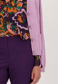 Lila geribbelde cardigan over een patroon blouse met oranje, groene en paarse ontwerpen, gecombineerd met passend paarse broeken.