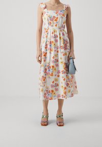 Robe midi florale en crème avec des fleurs colorées ; bretelles froncées, corsage ajusté et jupe fluide. Portée avec des sandales à talons multicolores.