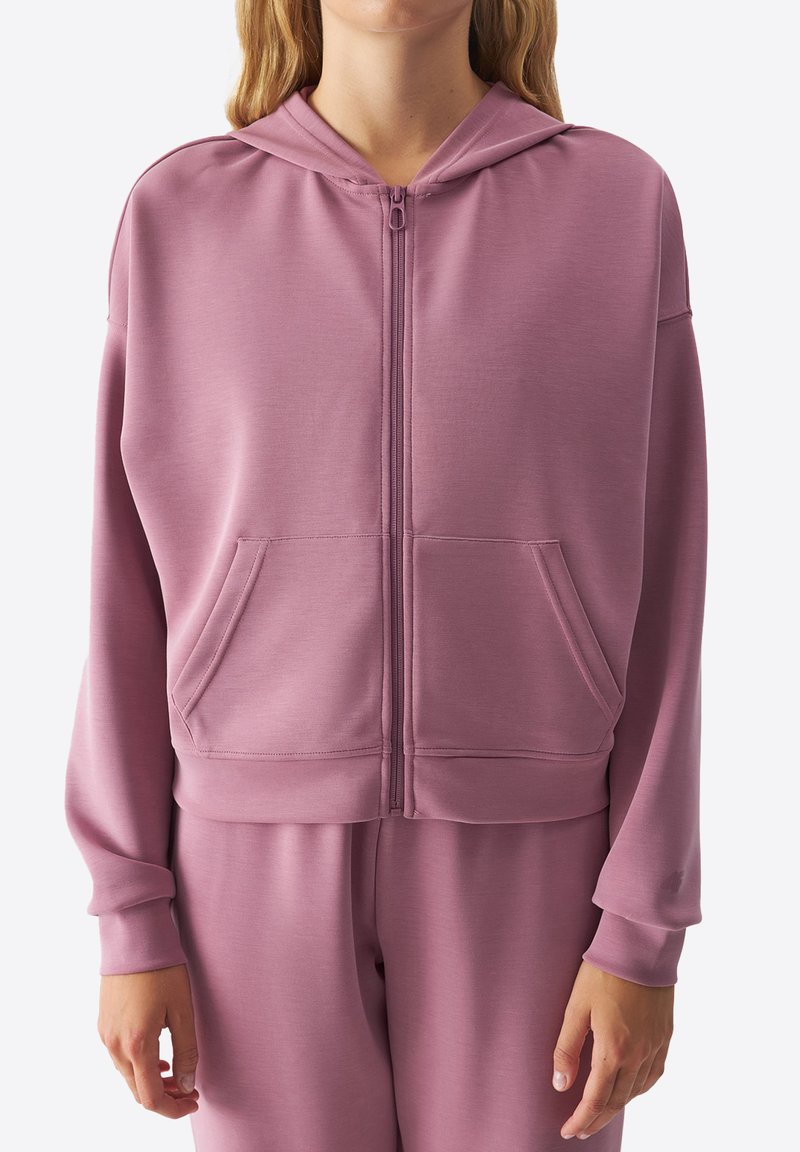 Sudadera con cremallera de color rosa claro, con cintura ajustada, mangas largas y dos bolsillos frontales. Textura suave, diseño minimalista y capucha.
