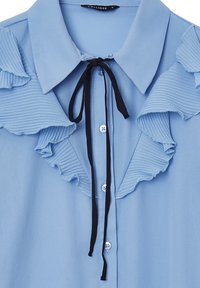 Blusa azzurra cielo realizzata in tessuto liscio, con dettagli arricciati lungo il colletto, una cravatta nera e chiusure a bottoni.