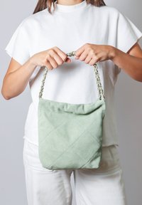 Borsa in suede verde trapuntato con manico a catena, caratterizzata da una texture morbida e una forma rettangolare, tenuta da una persona in un outfit bianco.