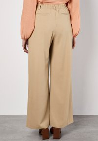 Femme portant un pantalon large beige avec une taille élastique et des chaussures block heel marron, se tenant devant un mur uni.