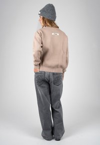 Femme portant un bonnet en maille gris, un pull beige avec étiquette et un jean gris ample, debout de dos sur un fond uni clair.