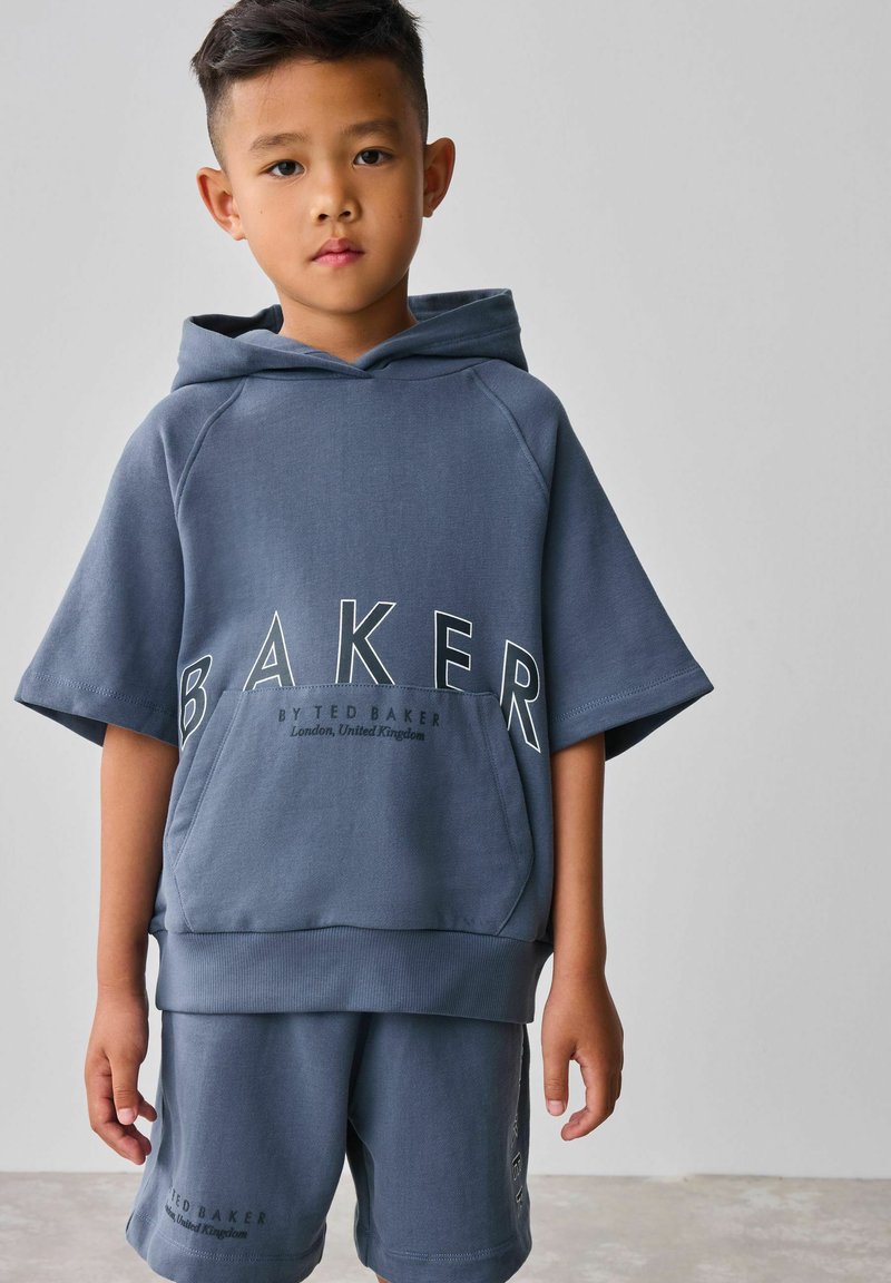 Modrý set mikiny s kapucí a šortek s velkým logem "BAKER" na přední straně, vyrobený z měkkého materiálu s volnějším střihem a krátkými rukávy.