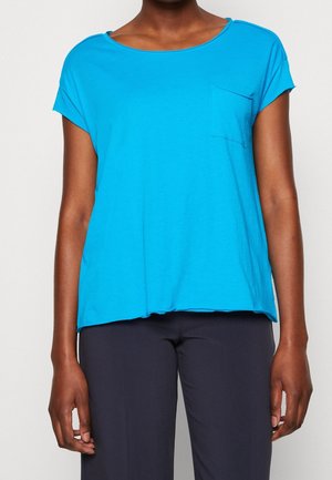 Frau trägt ein hellblaues Kurzarmoberteil mit einer kleinen Brusttasche und dunkelblauer Hose, dargestellt vom Hals bis zur Hüfte.