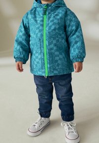 Giacca per bambini turchese con motivi di dinosauri, chiusura frontale con zip e dettagli verdi. Abbinata a jeans blu scuro e sneakers bianche.