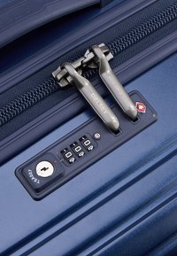Maleta azul marino con una superficie texturizada, que cuenta con un cierre doble, candado de combinación, orificio para llave y el logo de la marca "DELSY" en el tirador del zipper.