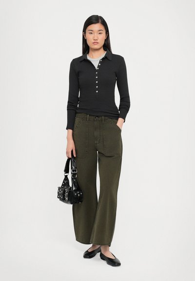 Zwarte geribbelde langärmelige top met knoopsluiting, gecombineerd met losse olijfgroene wijde broek, en een kleine zwarte tas met studs.