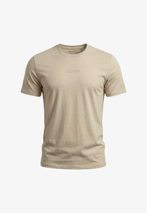 Camiseta beige de manga corta con cuello redondo y "GUESS" impreso en un sutil color a juego en el pecho, fondo liso.