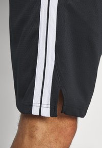 Des shorts de sport noirs en tissu mesh, avec des fentes sur les côtés et deux bandes verticales blanches de chaque côté. Surface texturée pour la respirabilité.