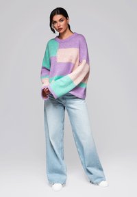 Un pull surdimensionné avec des blocs de couleur violet, vert et rose ; texture côtelée et manches larges ; associé à un jean large bleu clair et des baskets blanches.