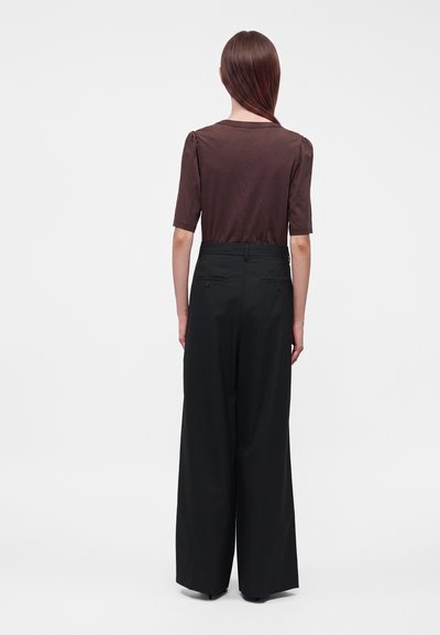 Femme aux cheveux bruns et raides, portant un haut marron à manches courtes et un pantalon noir à jambes larges, debout dos à l’observateur.