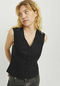 Crna majica bez rukava s V-izrezom, koja ima pet dugmadi sprijeda, dva bočna džepa i kroj koji naglašava figuru.