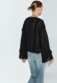 Svart, genomskinlig blus med volanger längs kanterna, med långa ärmar och lös passform, kombinerad med ljusblå denimjeans.