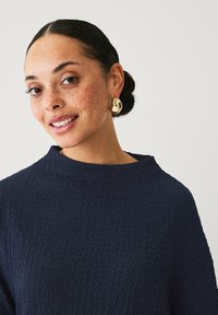 Mørkeblå struktureret top med høj hals, der har et tætsiddende design. Guld, rund ørering med et abstrakt udskæringsdetalje.