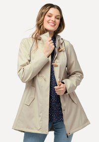Giacca impermeabile beige con cappuccio, chiusure a cordoncino e tasche laterali; indossata sopra una camicia blu a fantasia e abbinata a jeans.
