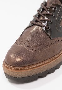 Chaussure oxford en tissu marron avec un dessus en cuir noir brillant, des détails perforés, une semelle intermédiaire texturée et une semelle extérieure noire robuste.