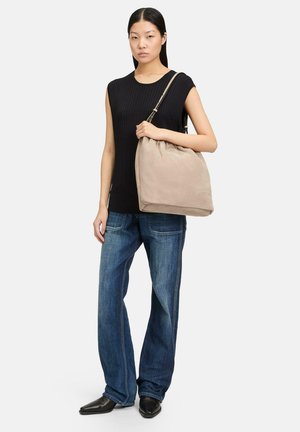 Bolso hobo de ante beige con cierre de cordón. El bolso presenta una textura suave y un logo grabado. La modelo lleva un top negro con canalé y jeans azules.