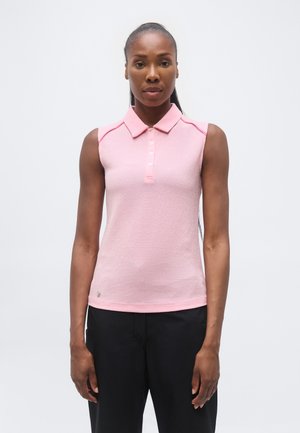 Frau mit dunkler Haut und langen Haaren trägt ein ärmelloses hellrosa Hemd mit Kragen und schwarze Hose, steht vor einem einfarbigen Hintergrund.