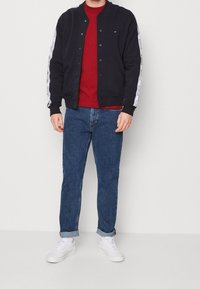 Marinblå bomberjacka med tryckknappar och vita sidostråk över en röd skjorta, kombinerad med blå jeans och vita sneakers.