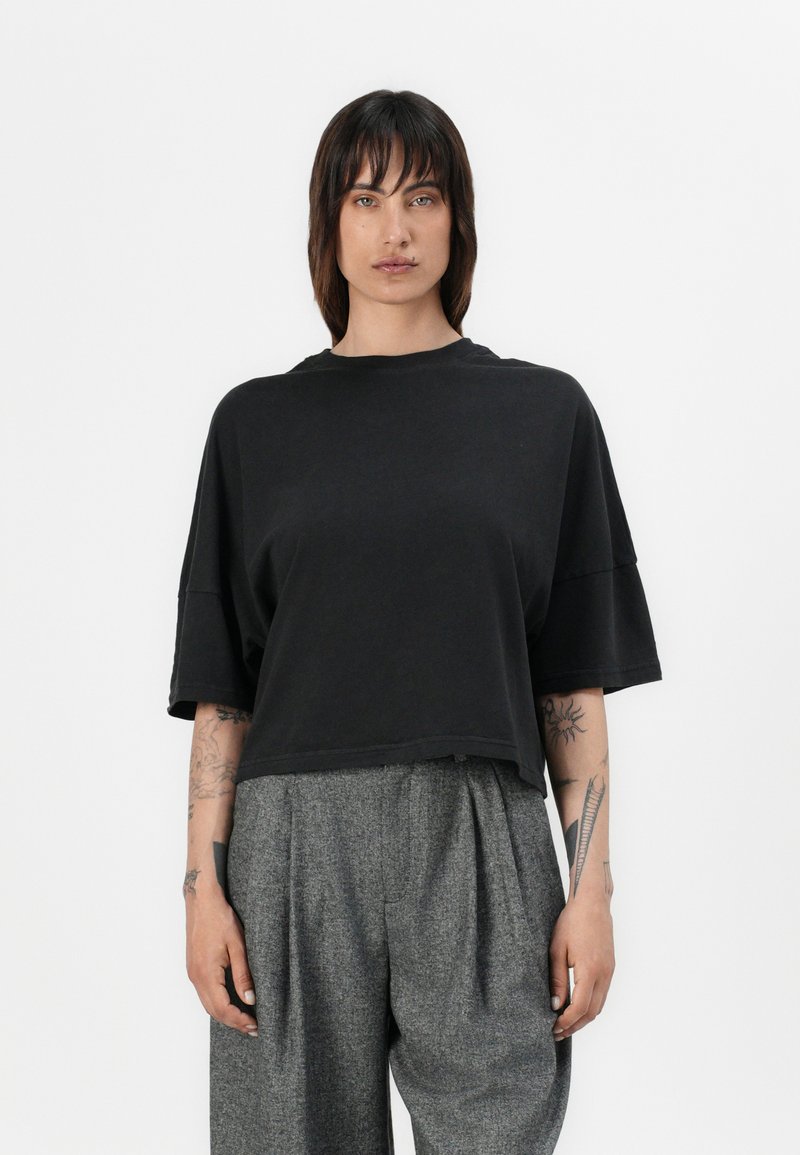 Svart kortermet t-skjorte med rund hals og brede korte ermer, kombinert med grå plissert bukse. Stoffet ser mykt og avslappet ut.
