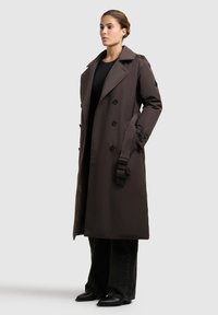 Donna in piedi, che indossa un lungo cappotto doppio petto marrone scuro con cintura e pantaloni neri a gamba larga, su uno sfondo grigio chiaro.