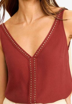 Femme portant un haut sans manches rouge rouille à encolure en V avec de petites clous métalliques ronds soulignant l'encolure et la couture centrale avant.